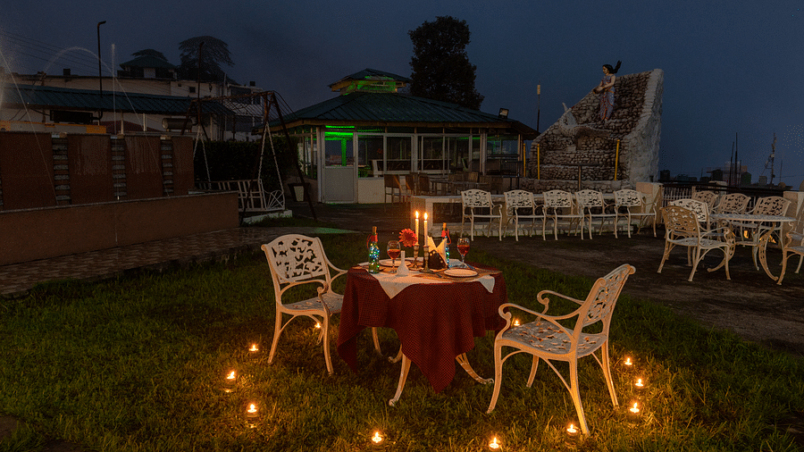 Candlelight dinner setup with a table for two, surrounded by candles and a view of a small fountain and seating area in the background - DLS Meghavan Resort, Dharamshala
